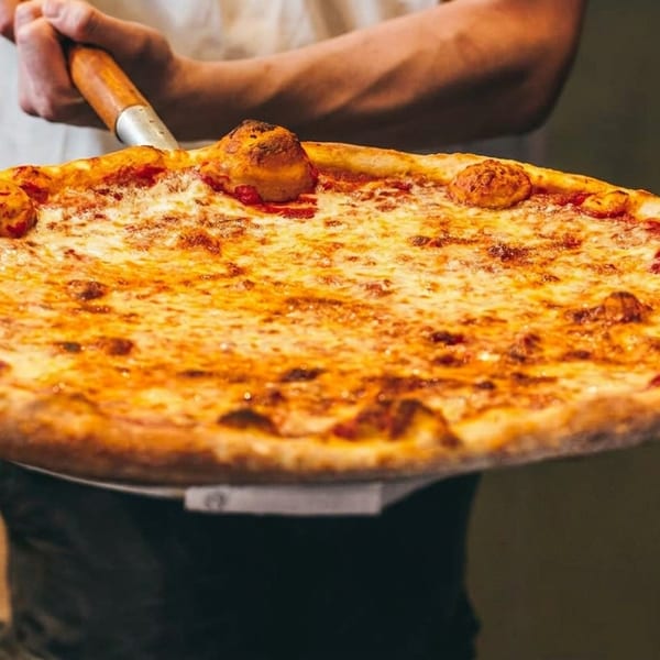 A large, hot cheese pizza being removed from a brick oven with a pizza peel.