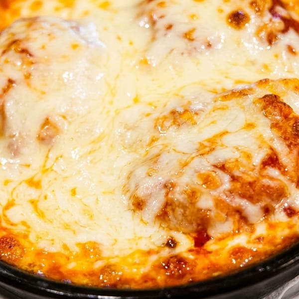 Close-up of baked meatballs covered in melted cheese and tomato sauce in a black dish.